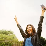 Vrouw wandelt in park en houdt beide armen omhoog met smartphone in één hand